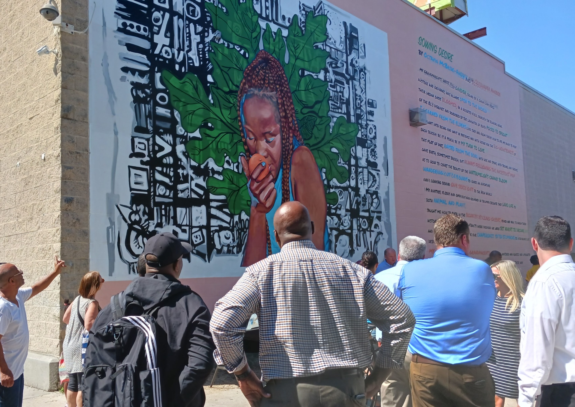 Visitors gather in front of a large outdoor mural featuring a figure holding a peach surrounded by green leaves and bold pattern